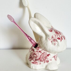 Peut inclure: Un porte-brosse à dents en céramique blanche en forme de lapin avec un motif floral rouge. Le support contient deux brosses à dents, une rose translucide et une blanche opaque, insérées dans le haut du dos et de la tête du lapin.