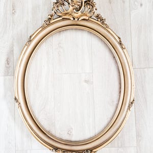 Peut inclure: Un cadre photo ovale doré orné de volutes en haut et en bas. Le cadre a un bord lisse et arrondi. Le cadre est vide, prêt pour une photo ou une œuvre d'art. Le cadre est fait d'un matériau doré.