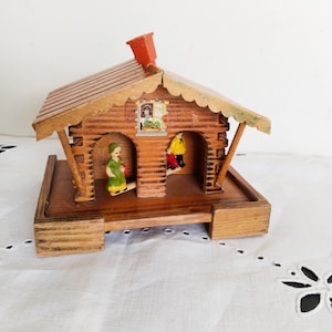 May include: A wooden cabin with a red roof and two small figures standing in the doorways. The cabin is on a wooden base and has a window with a painted scene.