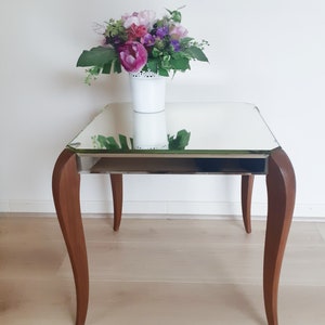Peut inclure: Une table d'appoint en miroir avec un cadre en bois et des pieds courbes. Un vase blanc avec des fleurs roses et violettes est posé sur la table.