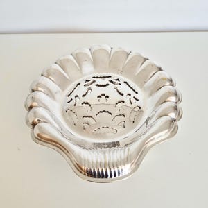 Peut inclure: Un plat en métal argenté en forme de coquille Saint-Jacques. Le plat présente un bord festonné et une zone circulaire centrale avec des motifs découpés symétriques et complexes qui ressemblent à des motifs floraux ou géométriques.