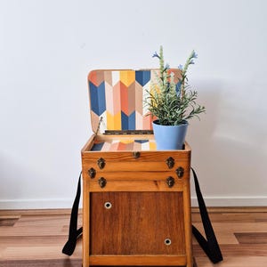 May include: A wooden sewing box with a hinged lid featuring a colorful geometric chevron pattern in orange, blue, and cream. The box has metal latches, black shoulder straps, and two circular metal eyelets on the front panel. A light blue pot with a green plant sits inside.