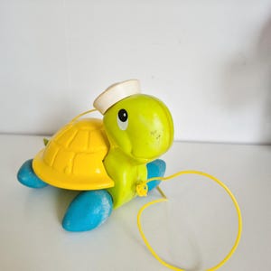 May include: A vintage pull-along toy turtle. The turtle has a yellow shell, green head, blue feet, and a white sailor hat. A yellow string is attached for pulling. The toy is made of plastic and is on a white surface.
