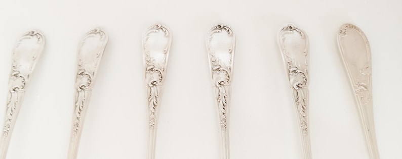 May include: Six silver spoons with intricate floral and scrollwork patterns. The spoons are displayed in a row against a white backdrop. The spoons have a classic, elegant appearance, suitable for formal dining or display.