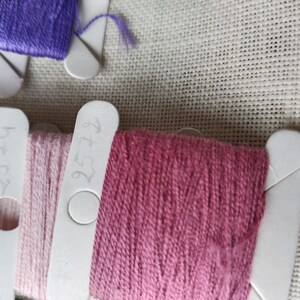 May include: Close-up of three spools of embroidery floss. The floss is wound around white plastic bobbins with numbers written on them. The numbers are 727, 375, and 476. The floss colors are pink, light pink, and a darker pink.