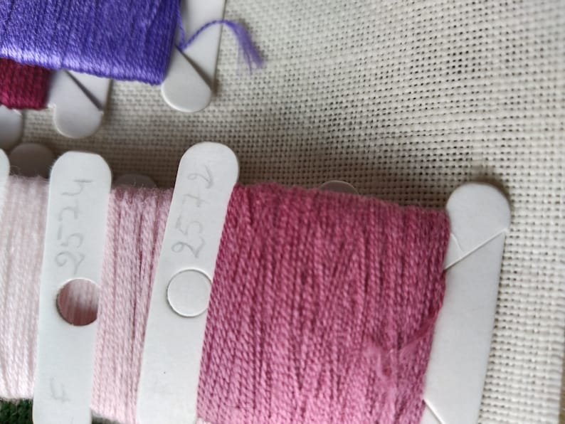 May include: Close-up of three spools of embroidery floss. The floss is wound around white plastic bobbins with numbers written on them. The numbers are 727, 375, and 476. The floss colors are pink, light pink, and a darker pink.