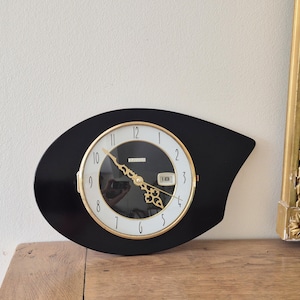 Peut inclure: Une horloge murale noire asymétrique avec un cadran circulaire blanc et des aiguilles dorées ornées. L'horloge présente des chiffres arabes noirs, une lunette dorée et une petite fenêtre de date rectangulaire affichant le chiffre 18.