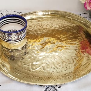 Puede incluir: Una bandeja grande y redonda de latón dorado con intrincados patrones florales en relieve. Un pequeño cuenco de cerámica con un diseño geométrico azul y blanco se encuentra en la bandeja. El cuenco tiene un borde azul y un interior blanco. La bandeja está sobre un mantel blanco.