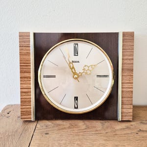 May include: A rectangular vintage clock with a cream-colored face and gold-tone hands and accents. The clock face has black markings and the word "BODYA". The clock is encased in a dark brown frame with wood-grain side panels.