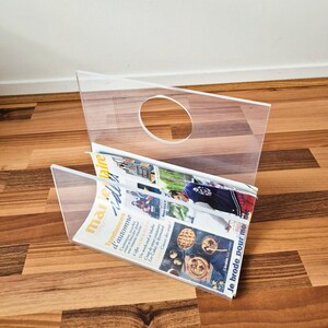 May include: A clear acrylic magazine rack with a central circular cutout handle. The rack is constructed from transparent panels and holds a magazine titled Marie Claire Idées with visible text and imagery on the cover, including autumn themes.