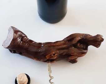 May include: A wooden corkscrew shaped like a tree branch with a metal worm. The corkscrew is lying on a white surface. A cork and a bottle of wine are also visible in the image.