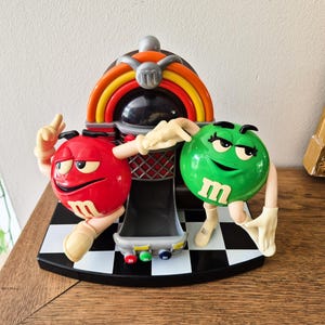 May include: A plastic M&M's candy dispenser shaped like a jukebox with a red character on the left and a green character on the right. The base features a black and white checkered pattern, and the jukebox has colorful arches and three buttons.