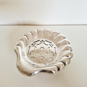 Könnte beinhalten: Eine silberfarbene, muschelförmige Schale mit gewelltem Rand und einem dekorativen, durchbrochenen Design in der Mitte. Die Schale hat eine griffartige Verlängerung mit vertikalen Rippen. Das Gesamtdesign ist verziert und vintage.