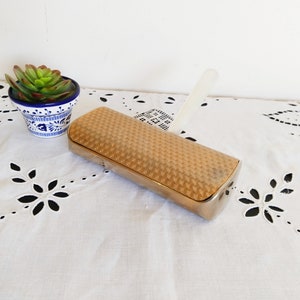 Peut inclure: Une boîte rectangulaire décorative de couleur dorée avec un motif tissé sur le dessus. La boîte est ouverte et possède une poignée en plastique blanc.
