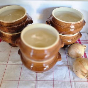 Peut inclure: Six bols à soupe en céramique marron avec des poignées, disposés sur une nappe à carreaux blancs et rouges. Deux oignons jaunes sont à côté des bols. Les bols ont un intérieur beige clair.
