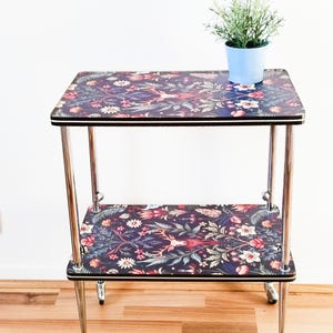 Può includere: Tavolino a due livelli con motivo floreale. Il tavolo ha uno sfondo scuro con un motivo di fiori, cervi e fogliame. Il telaio è cromato e il tavolo ha piccole ruote.