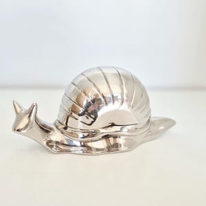 Peut inclure: Une figurine d'escargot argentée avec une coquille striée et un corps détaillé. La pièce décorative est en métal et repose sur une surface blanche. Le corps de l'escargot est lisse et brillant.