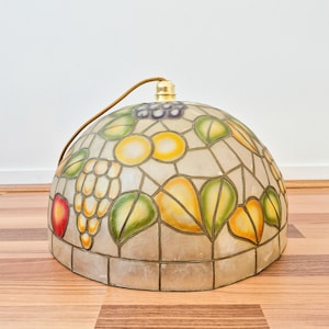 May include: A dome-shaped stained glass lamp shade featuring a fruit motif with grapes, apples, and pears in shades of red, yellow, and green. The glass panels are held together by dark metal lines, and a gold-toned metal fixture sits at the top.