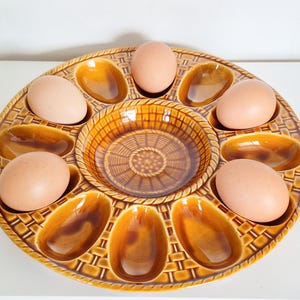 Puede incluir: Una bandeja redonda de cerámica esmaltada en color marrón con una textura de tejido de cesta. La bandeja tiene un cuenco circular central rodeado por ocho hendiduras ovaladas, con cuatro huevos de color marrón claro en los huecos.