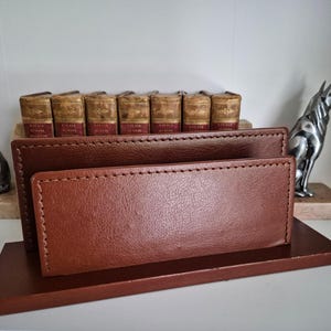 Könnte beinhalten: Ein brauner Leder-Schreibtisch-Organizer mit rechteckiger Basis und zwei Abschnitten. Der Organizer ist von dekorativen Gegenständen umgeben, darunter kleine Bücher und silberne Hundestatuen. Das Leder hat sichtbare Nähte.