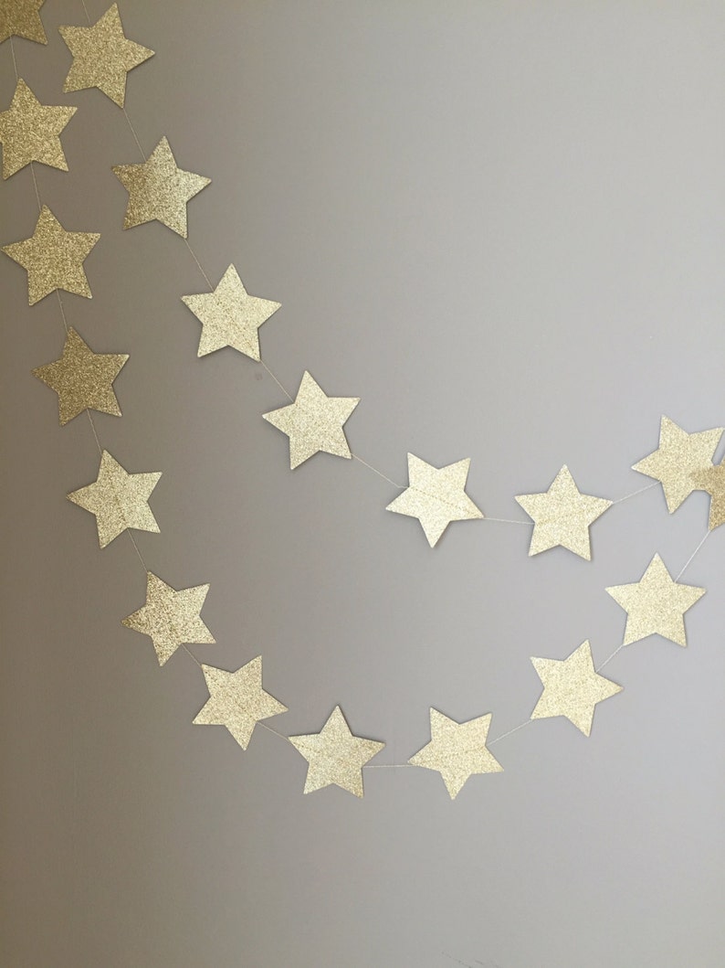 May include: A gold glitter star garland with a string of stars hanging in a curved shape against a light gray wall.