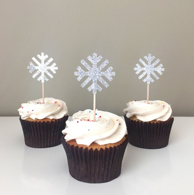 Puede incluir: Tres cupcakes con glaseado blanco y chispas de colores coronados con adornos de cupcake de copo de nieve plateado con purpurina.