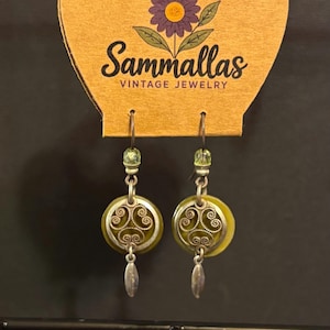 May include: A pair of dangle earrings featuring olive green circular discs with silver-toned filigree overlays, small light green beads, and silver-toned leaf-shaped charms. The earrings are attached to a tan card labeled Sammallas Vintage Jewelry.