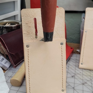 May include: A handmade leather tool holder with a wooden handle tool. The holder is light tan with a stitched border and metal rivets. The tool handle is brown with a black base and purple paint on the top.