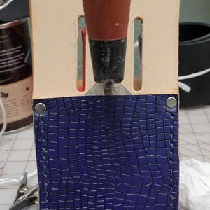 May include: A leathercraft tool with a brown handle is inserted into a blue leather sheath. The sheath has a textured, crocodile-like pattern. The top portion of the sheath is a light tan color. The tool is designed for leatherworking.