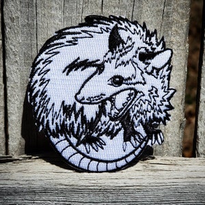 May include: Embroidered patch featuring a stylized opossum in a curled position. The design uses black thread on a white background, showcasing the animal's features and fur texture. The patch is circular and has a black border.