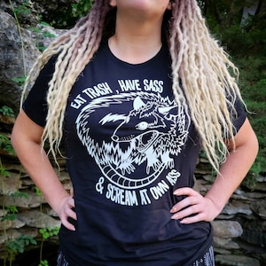 May include: Black t-shirt with a white graphic of a possum with the text "Eat Trash, Have Sass & Scream At Own Ass".