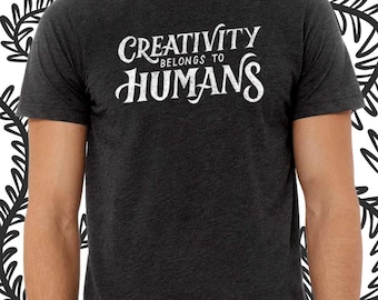 Könnte beinhalten: Dunkelgraues T-Shirt mit dem weißen Text "CREATIVITY BELONGS TO HUMANS". Das kurzärmlige Shirt hat einen Rundhalsausschnitt. Der Text ist in einer stilisierten Schriftart gehalten.