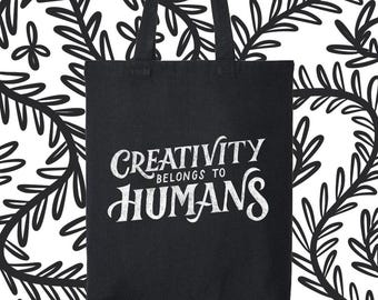 Könnte beinhalten: Schwarze Tragetasche mit dem weißen Text "CREATIVITY BELONGS TO HUMANS". Die Tasche hat zwei schwarze Griffe und befindet sich vor einem weißen Hintergrund mit einem schwarzen Blättermuster.