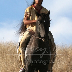 Puede incluir: Un hombre nativo americano con ropa tradicional monta un caballo negro con una estrella blanca en la frente. Lleva un collar de cuentas y una pluma en el pelo. El caballo lleva una brida y una silla de montar de cuero. El fondo es un campo de hierba alta.