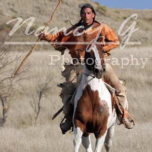 Puede incluir: Un hombre con ropa tradicional monta un caballo marrón y blanco mientras sostiene una lanza de madera. Lleva un chaleco de cuero marrón y una camisa larga de color naranja. El caballo corre por un campo de hierba alta.