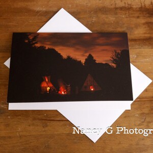 May include: A nighttime scene with three lit tents in a wooded area. The sky is a vibrant orange and pink with clouds.