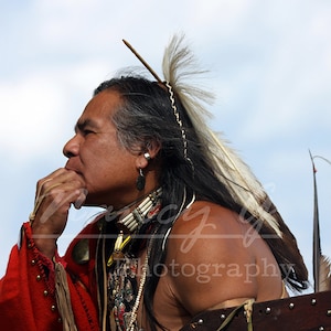Puede incluir: Un hombre nativo americano con vestimenta tradicional, visto de perfil contra un cielo nublado. Lleva una prenda roja, collares de cuentas y un tocado de plumas. Su cabello oscuro está adornado con plumas y un alfiler de madera.