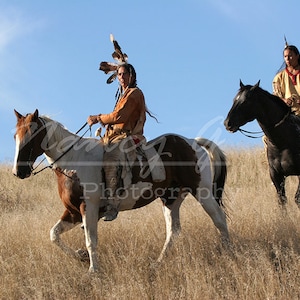 Puede incluir: Dos personas con vestimenta tradicional nativa americana montan a caballo por un campo de hierba. La persona de la izquierda lleva un tocado de plumas y la persona de la derecha lleva un chaleco con cuentas. Ambos caballos son marrones y blancos.