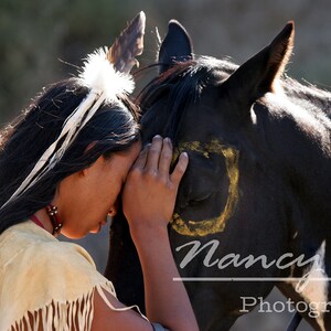 Puede incluir: Una persona joven con cabello largo y oscuro que lleva un atuendo tradicional nativo americano con plumas y flecos, toca suavemente la cara de un caballo negro con una marca amarilla. El caballo tiene los ojos cerrados y parece estar tranquilo.