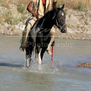 Puede incluir: Un hombre nativo americano con vestimenta tradicional monta un caballo negro y blanco a través de un río poco profundo. El caballo está salpicando agua mientras camina por el agua.
