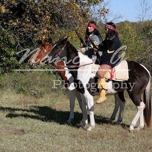 Puede incluir: Dos personas a caballo con vestimenta y tocado tradicional nativo americano. Están cabalgando por un campo de hierba con árboles en el fondo.