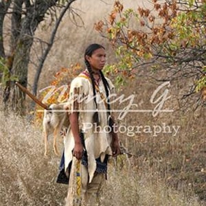 Puede incluir: Un hombre nativo americano con ropa tradicional y llevando una lanza se encuentra en un campo de hierba alta. Lleva una camisa y pantalones de piel de venado blancos, y su largo cabello negro está trenzado. El fondo es una imagen borrosa de árboles y arbustos.