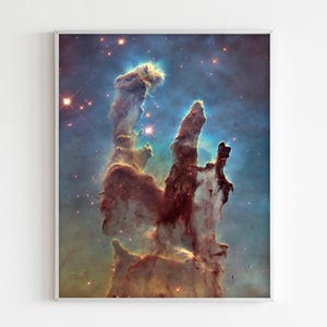 May include: A framed print featuring the Pillars of Creation, a vibrant nebula in space. The image showcases towering columns of gas and dust in shades of brown, gold, and blue, with numerous stars scattered throughout the scene.