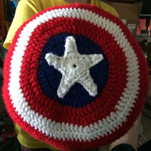 May include: A child holds a crocheted red, white, and blue Captain America shield. The shield has a white star in the center on a blue background.