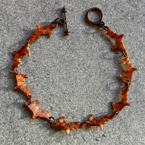 May include: A copper-toned bracelet with a unique design. The bracelet features a series of jagged, organic shapes linked together. It has a toggle clasp closure. The metal has a textured, weathered appearance, giving it a rustic aesthetic.