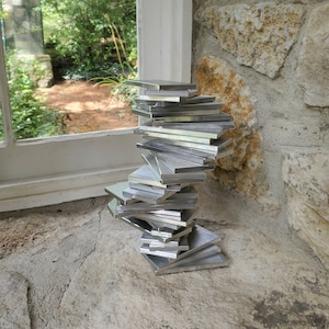 Könnte beinhalten: Ein Stapel silberner Metallfliesen, in Form eines schiefen Turms angeordnet, steht auf einer rauen, grauen Steinoberfläche. Die Fliesen sind versetzt gestapelt und bilden eine dynamische und abstrakte Skulptur.