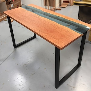 May include: A wooden table with a black metal frame. The table top is made of wood with a clear epoxy river running through the center. The table is in a workshop setting with tools and other wood pieces in the background.