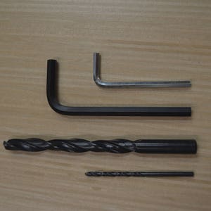 May include: Four tools on a wooden surface: a black L-shaped Allen key, a silver L-shaped Allen key, a black drill bit, and a smaller black drill bit.
