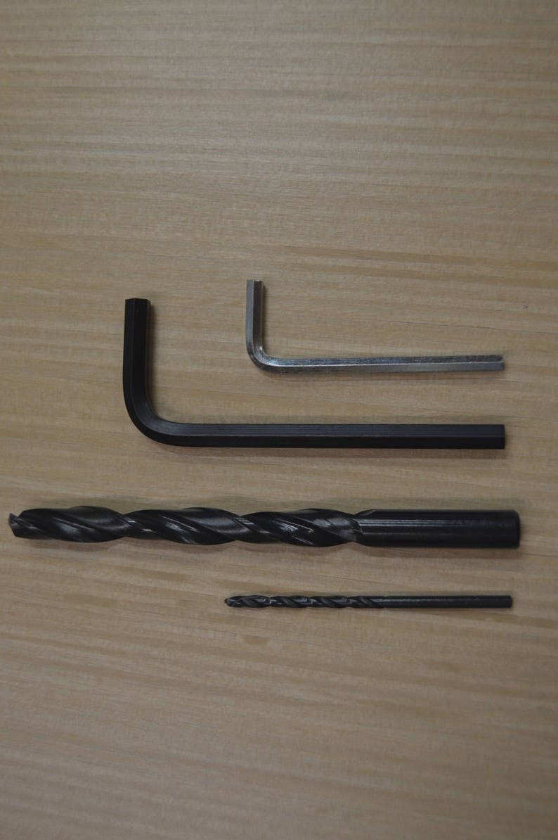 May include: Four tools on a wooden surface: a black L-shaped Allen key, a silver L-shaped Allen key, a black drill bit, and a smaller black drill bit.