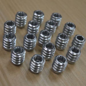 May include: A close-up of a group of silver metal threaded inserts. The inserts are arranged in a circular pattern on a wooden surface.